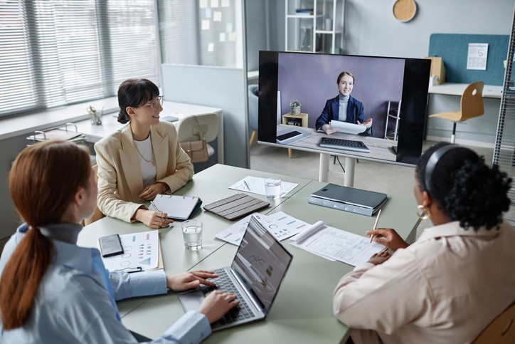 High angle shot of female business colleagues sitting at table in modern office and looking at plasma screen while having online video call meeting