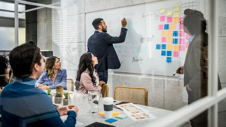 Diverse coworkers writing on whiteboard and generating ideas in conference room. Multiracial business people using agile methodology for project planning, view behind window