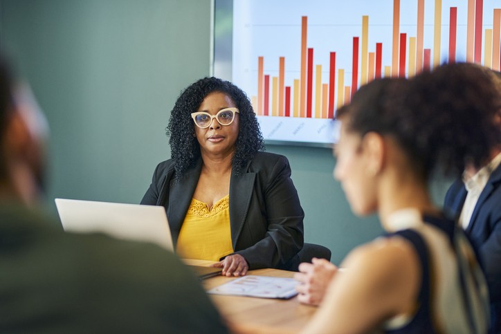 A confident businesswoman wearing glasses leads a meeting with her colleagues in a modern office. They are discussing the company's financial performance, which is displayed on a screen behind them.