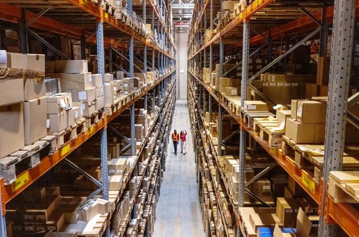 High angle view of a warehouse manager walking with foremen checking stock on racks. Businesswoman discussing stock with a male worker while walking by racks in warehouse.