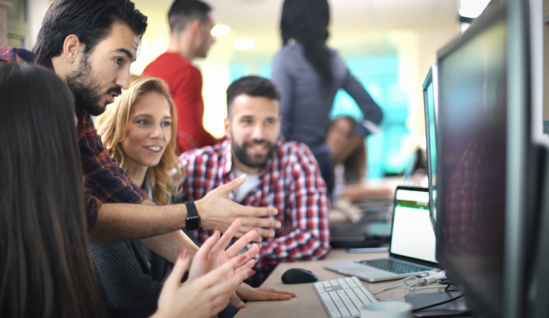 Closeup side view of group of application developers discussing about certain code for an application. There are two men and two women standing and sitting in front of a computer, one of them is explaining something about the code on the screen.. B;urry people in background, also released.