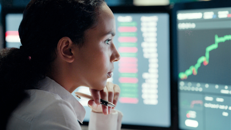 Young corporate businesswoman or Financial Analyst reading and studying investment charts on multiple computer screens. Confident female analyzing future stock market data sitting down.